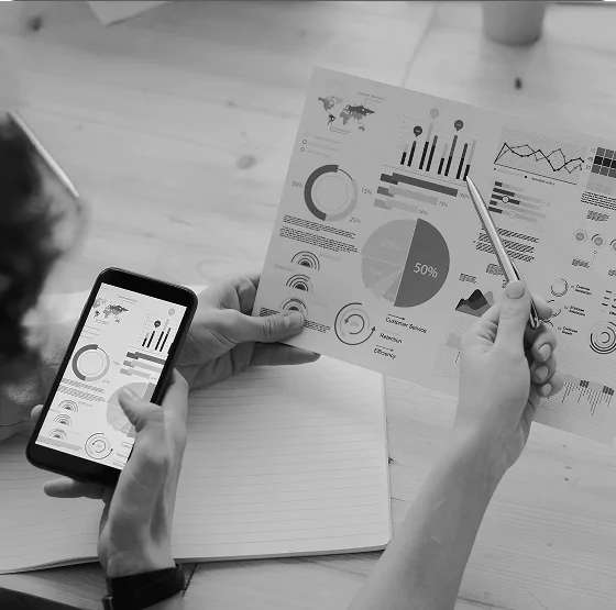 Two people analyzing charts and graphs on a smartphone and printed paper, illustrating data analytics and performance reporting.